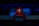 A person wearing a red hoodie works on a laptop in a dark room illuminated by blue light. The background features server equipment and logos of various technology companies on a transparent panel.