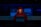 A person wearing a red hoodie works on a laptop in a dark room illuminated by blue light. The background features server equipment and logos of various technology companies on a transparent panel.