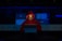 A person wearing a red hoodie works on a laptop in a dark room illuminated by blue light. The background features server equipment and logos of various technology companies on a transparent panel.