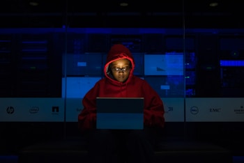 A person wearing a red hoodie works on a laptop in a dark room illuminated by blue light. The background features server equipment and logos of various technology companies on a transparent panel.