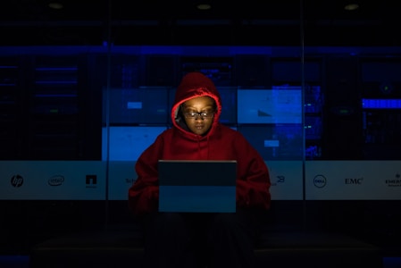 A person wearing a red hoodie works on a laptop in a dark room illuminated by blue light. The background features server equipment and logos of various technology companies on a transparent panel.