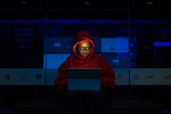 A person wearing a red hoodie works on a laptop in a dark room illuminated by blue light. The background features server equipment and logos of various technology companies on a transparent panel.