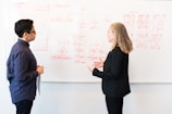 Two people engage in a discussion in front of a whiteboard filled with diagrams and flowcharts. One person is holding a red pen and a sheet of paper, while the other gestures with their hands, possibly explaining or elaborating on the ideas.