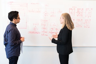 Two people engage in a discussion in front of a whiteboard filled with diagrams and flowcharts. One person is holding a red pen and a sheet of paper, while the other gestures with their hands, possibly explaining or elaborating on the ideas.