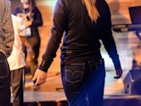 A group of people are gathered indoors, with some focusing on a stage area where musical equipment is present. The lighting is warm, creating a casual and relaxed atmosphere.