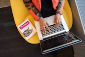 A person is seated on a yellow chair, working on a laptop with visible code on the screen. Next to them, a book titled 'Python' is placed.