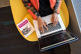 A person is seated on a yellow chair, working on a laptop with visible code on the screen. Next to them, a book titled 'Python' is placed.