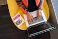 A person is seated on a yellow chair, working on a laptop with visible code on the screen. Next to them, a book titled 'Python' is placed.