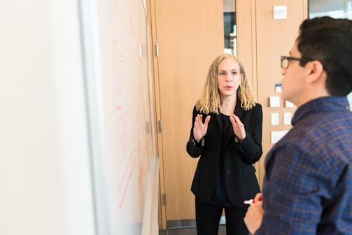 A woman with curly blonde hair, dressed in a black suit, is actively explaining something in front of a whiteboard filled with red marker sketches. A person wearing a plaid shirt is seen from the back, holding a red marker and facing the woman, indicating active engagement in the discussion.