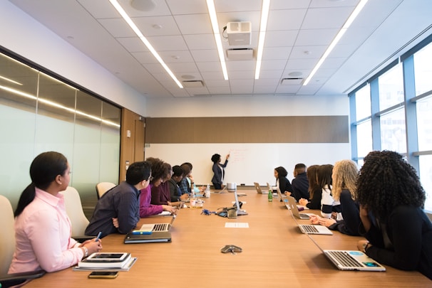 A group of people seated around a long conference table, attentively listening to someone presenting information on a whiteboard. The setting is a modern conference room with large windows that let in natural light. Various individuals have laptops open in front of them, and there are bottles of water on the table. The atmosphere is professional and focused.