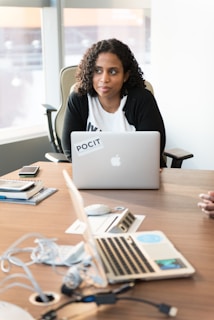 A person with curly hair sits at a desk in an office environment, looking to the side, with a laptop in front of them. The table is scattered with electronic devices, documents, and personal items, indicating a workspace.