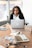 Photo of a thoughtful woman with curly hair, sitting at a desk with a laptop.