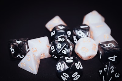 A collection of polyhedral dice with various numbers of sides, featuring black and white surfaces with numbers in contrasting colors. The dice are scattered on a dark background, displaying numbers in a mix of white and orange.