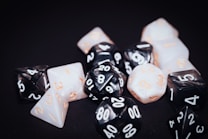 A collection of polyhedral dice with various numbers of sides, featuring black and white surfaces with numbers in contrasting colors. The dice are scattered on a dark background, displaying numbers in a mix of white and orange.