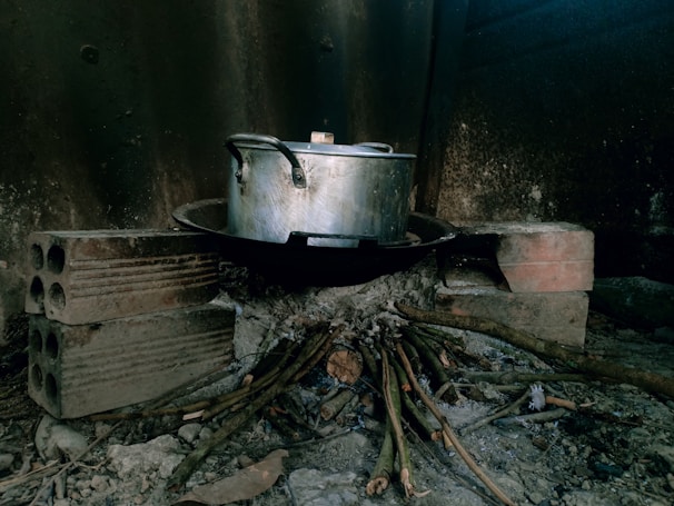 A metal pot with a handle sits on top of a makeshift stove constructed from bricks. Beneath the pot, a metal griddle holds up the pot. Small sticks and twigs are gathered underneath the stove, arranged to serve as fuel for a fire. The background is a dark, rustic surface, possibly a wall.