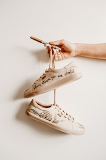 A hand holding a pair of beige sneakers that are creatively personalized with floral designs and the phrase 'bloom where you are planted'. The sneakers hang on a beige background, giving a minimalist and artistic vibe.
