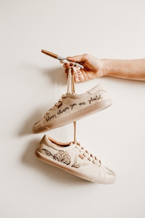 A hand holding a pair of beige sneakers that are creatively personalized with floral designs and the phrase 'bloom where you are planted'. The sneakers hang on a beige background, giving a minimalist and artistic vibe.