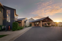 A modern church building surrounded by a landscaped garden and path. The architecture includes brick walls, a large entrance with a prominent roof, and a cross visible on top. The sky is partly cloudy with a sunset in the background creating a warm glow.