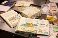 An arrangement of open illustrated books with colorful and artistic designs on the pages, placed on a table alongside art supplies like ribbons and containers. The pages contain text with vibrant illustrations including flowers and a sun.
