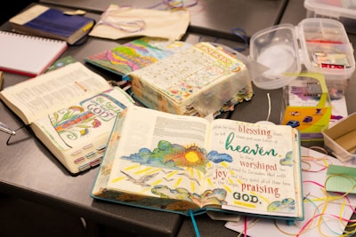 An arrangement of open illustrated books with colorful and artistic designs on the pages, placed on a table alongside art supplies like ribbons and containers. The pages contain text with vibrant illustrations including flowers and a sun.