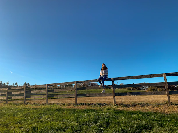 A happy customer wearing a Rustic Thread Co t-shirt while sitting on a wooden fence in a sunlit field.