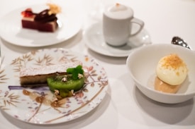 An elegant dessert spread featuring a slice of cake dusted with powdered sugar and a slice of kiwi on a floral plate, a bowl of creamy dessert topped with golden crumbs, and a small cup of cappuccino with frothy milk on top.
