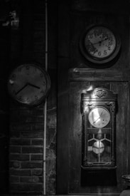 Three clocks are mounted on a wall, each with a distinct style. The clock on the left has a simple design with a white face and black numerals. The upper right clock appears older, showing signs of wear, with faded numerals and hands. Below it, a pendulum clock is encased in a vintage wooden frame, displaying intricate details.