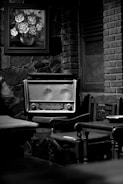 A vintage radio sits on a wooden chair surrounded by wooden furniture. A framed painting of flowers in a vase hangs on a stone wall behind it. The setting has an old-fashioned, rustic feel with elements of stone and brick.