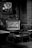 A vintage radio sits on a wooden chair surrounded by wooden furniture. A framed painting of flowers in a vase hangs on a stone wall behind it. The setting has an old-fashioned, rustic feel with elements of stone and brick.