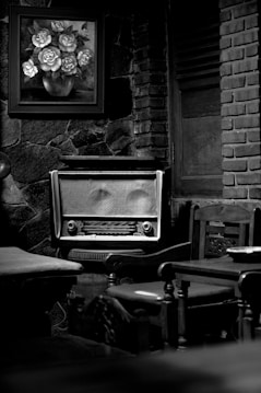 A vintage radio sits on a wooden chair surrounded by wooden furniture. A framed painting of flowers in a vase hangs on a stone wall behind it. The setting has an old-fashioned, rustic feel with elements of stone and brick.