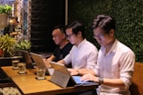 Three individuals are sitting at a wooden table in a work or study setting, each focused on their laptops. The background features a wall covered with greenery, creating a natural ambiance. Glasses of water are placed in front of them, and a potted plant can be seen in the corner.