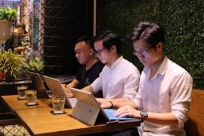 Three individuals are sitting at a wooden table in a work or study setting, each focused on their laptops. The background features a wall covered with greenery, creating a natural ambiance. Glasses of water are placed in front of them, and a potted plant can be seen in the corner.