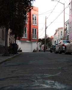 An urban street scene features a perspective view of a residential area. On the left, a modern, multi-story building with a red facade stands out. The building has large windows and a garage door at ground level. Several cars are parked along the street, and there are power lines above. Trees and shrubs are visible near the buildings.