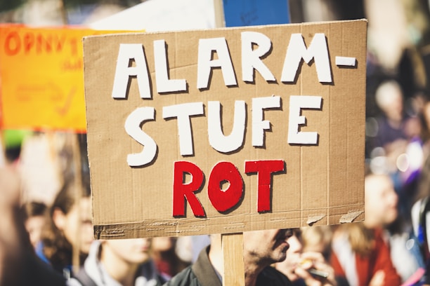 A close-up view of a protest sign made of cardboard with bold, white and red lettering. The words 'ALARMSTUFE ROT' are prominently displayed, with the bottom word emphasized in red. In the background, a crowd of people is visible, slightly out of focus, suggesting a protest or public gathering setting.