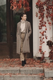 A person is wearing a long checkered coat and stands on steps covered with fallen autumn leaves. Red vines with leaves drape around a dark wooden door, creating a cozy and seasonal ambiance. The person appears relaxed and contemplative.
