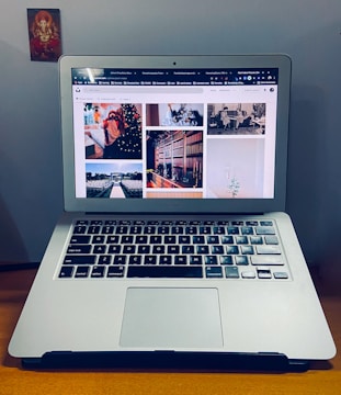 A laptop with an open screen displaying multiple photos, placed on a wooden surface. To the left is a small religious image stuck on the wall.