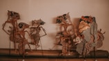 A collection of traditional shadow puppets arranged on a display surface. The puppets are intricately designed with detailed patterns and are made from thin materials that cast shadows when illuminated. The puppets represent various characters with exaggerated features, dressed in ornate costumes with elaborate headgear.