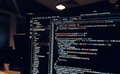 A computer monitor displays a Visual Studio Code interface with a dark theme. The screen shows lines of code written in JavaScript or HTML with syntax highlighting. The code includes functions, variables, and elements typical of web development. The background reveals a dimly lit office environment.