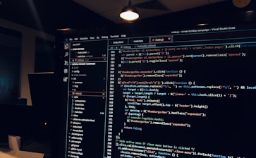 A computer monitor displays a Visual Studio Code interface with a dark theme. The screen shows lines of code written in JavaScript or HTML with syntax highlighting. The code includes functions, variables, and elements typical of web development. The background reveals a dimly lit office environment.