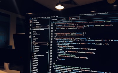 A computer monitor displays a Visual Studio Code interface with a dark theme. The screen shows lines of code written in JavaScript or HTML with syntax highlighting. The code includes functions, variables, and elements typical of web development. The background reveals a dimly lit office environment.