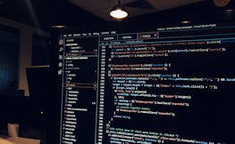 A computer monitor displays a Visual Studio Code interface with a dark theme. The screen shows lines of code written in JavaScript or HTML with syntax highlighting. The code includes functions, variables, and elements typical of web development. The background reveals a dimly lit office environment.