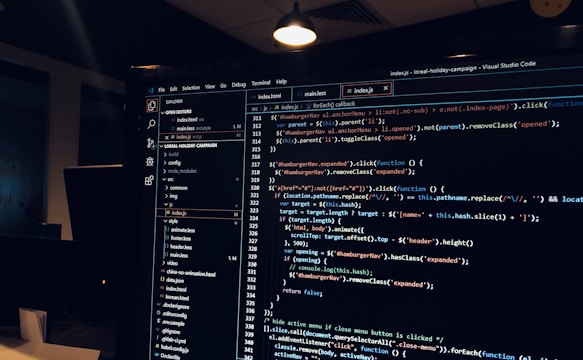 A computer monitor displays a Visual Studio Code interface with a dark theme. The screen shows lines of code written in JavaScript or HTML with syntax highlighting. The code includes functions, variables, and elements typical of web development. The background reveals a dimly lit office environment.