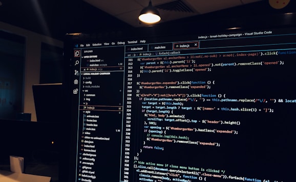 A computer monitor displays a Visual Studio Code interface with a dark theme. The screen shows lines of code written in JavaScript or HTML with syntax highlighting. The code includes functions, variables, and elements typical of web development. The background reveals a dimly lit office environment.