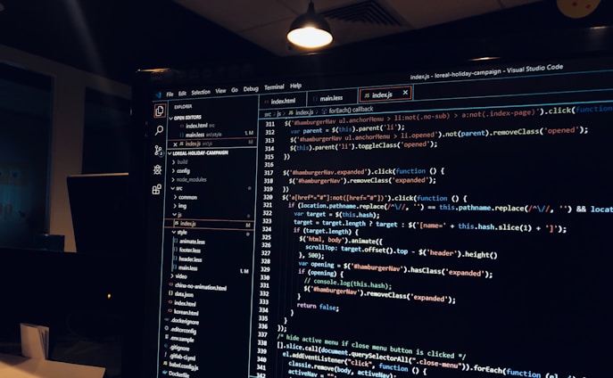 A computer monitor displays a Visual Studio Code interface with a dark theme. The screen shows lines of code written in JavaScript or HTML with syntax highlighting. The code includes functions, variables, and elements typical of web development. The background reveals a dimly lit office environment.