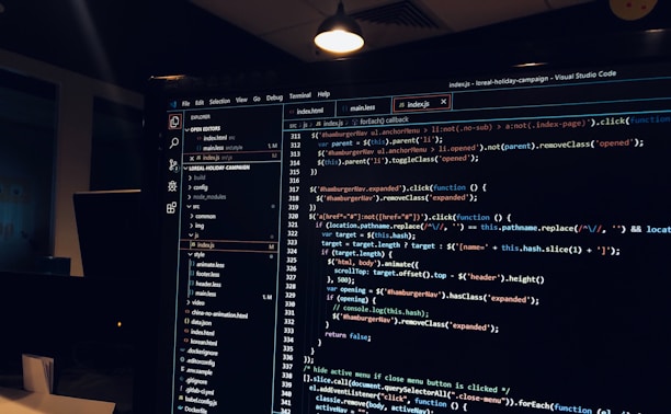 A computer monitor displays a Visual Studio Code interface with a dark theme. The screen shows lines of code written in JavaScript or HTML with syntax highlighting. The code includes functions, variables, and elements typical of web development. The background reveals a dimly lit office environment.