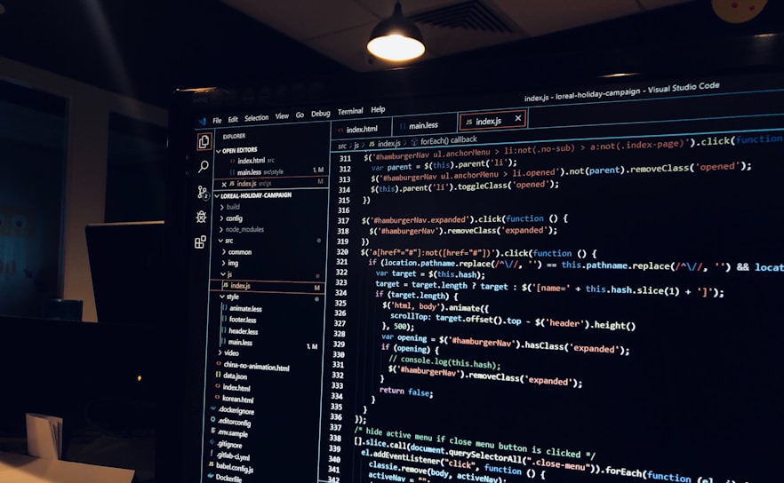 A computer monitor displays a Visual Studio Code interface with a dark theme. The screen shows lines of code written in JavaScript or HTML with syntax highlighting. The code includes functions, variables, and elements typical of web development. The background reveals a dimly lit office environment.