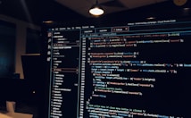 A computer monitor displays a Visual Studio Code interface with a dark theme. The screen shows lines of code written in JavaScript or HTML with syntax highlighting. The code includes functions, variables, and elements typical of web development. The background reveals a dimly lit office environment.