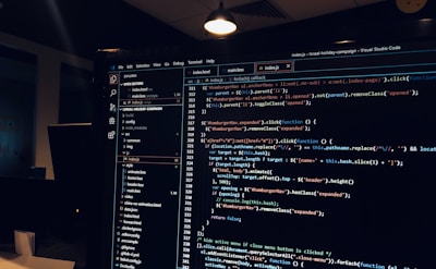 A computer monitor displays a Visual Studio Code interface with a dark theme. The screen shows lines of code written in JavaScript or HTML with syntax highlighting. The code includes functions, variables, and elements typical of web development. The background reveals a dimly lit office environment.