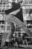 A large black and white advertisement on a glass building facade featuring a figure in denim jeans and a black jacket. The building reflects a city street with pedestrians visible through the glass. There are brand names, like 'Pro', partially visible within the advertisement.