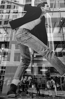 A large black and white advertisement on a glass building facade featuring a figure in denim jeans and a black jacket. The building reflects a city street with pedestrians visible through the glass. There are brand names, like 'Pro', partially visible within the advertisement.